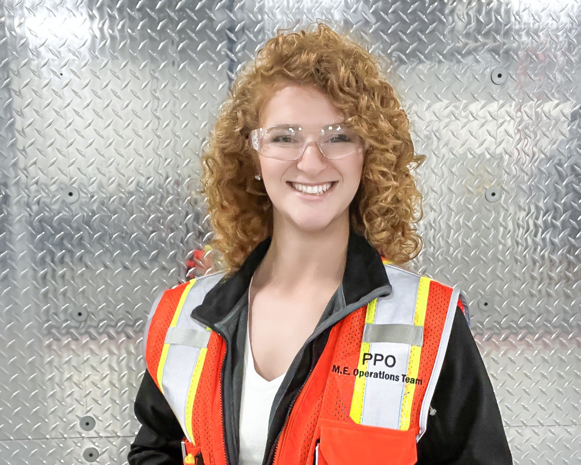A young woman with red hair wearing safety glasses and an orange safety vest. She is smiling in front of a metal wall.
