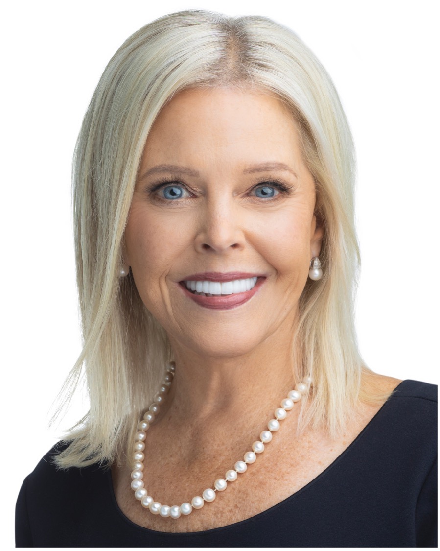 A smiling woman with blonde hair and blue eyes against a white background. She is wearing a black top, pearl necklace, and pearl earrings.