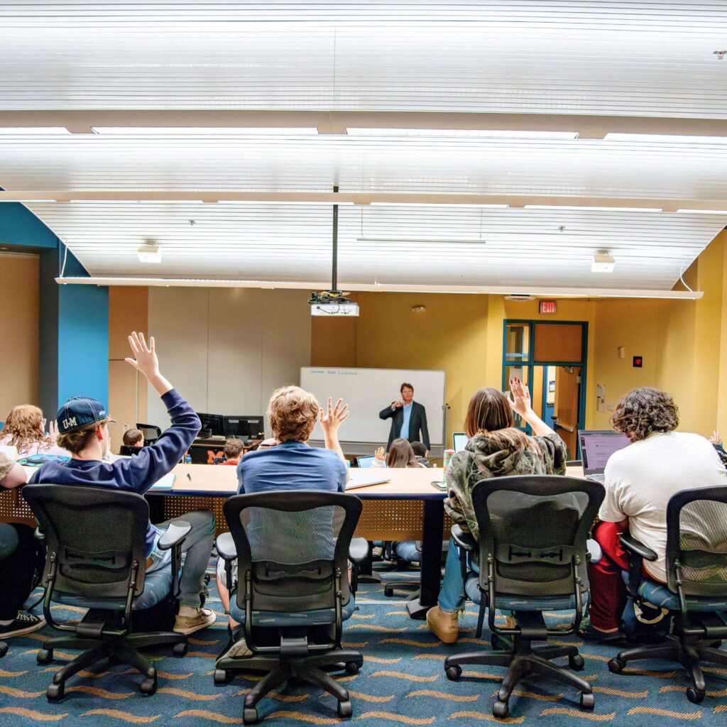 A classroom scene with several students raising their hands. A teacher at the front of the room gestures as if to call on a student.