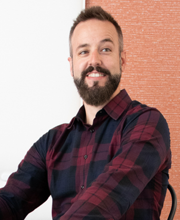 A portrait of a smiling man with a dark, full beard and short hair. He is wearing a plaid, button-down shirt that is red and navy blue. The background is a vertical, woven tapestry that is light red on an off-white wall.