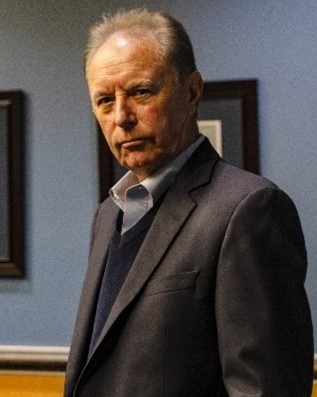 A man in a suit looks at the camera with his body turned to the left. The background features framed pictures on a blue wall.