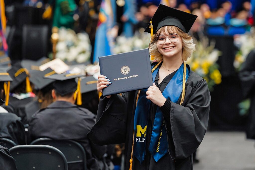 A young woman in a graduation cap and gown smiles while holding a diploma from the University of Michigan-Flint. A graduation ceremony is taking place in the background.