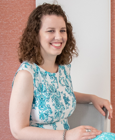 A seated portrait of a smiling woman with dark, curly hair. She is wearing a white, sleeveless dress that has an ornate turquoise and blue pattern. The background is a vertical, woven tapestry that is light red on an off-white wall.