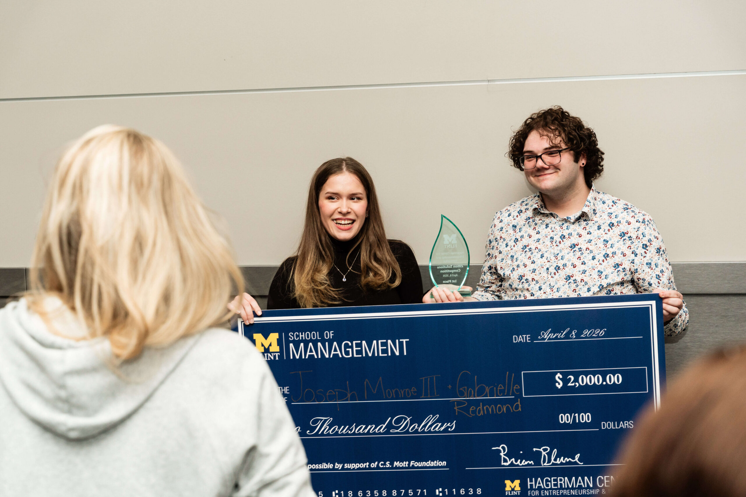 A young woman is on the left, and a young man is on the right. Together, they are holding a glass award and a large check for $2,000 from the School of Management.
