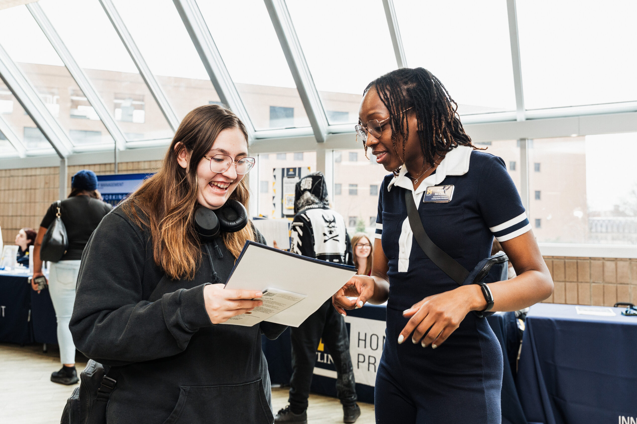 A person smiles while looking at a document, engaging with another person who is explaining something in a bright indoor setting.