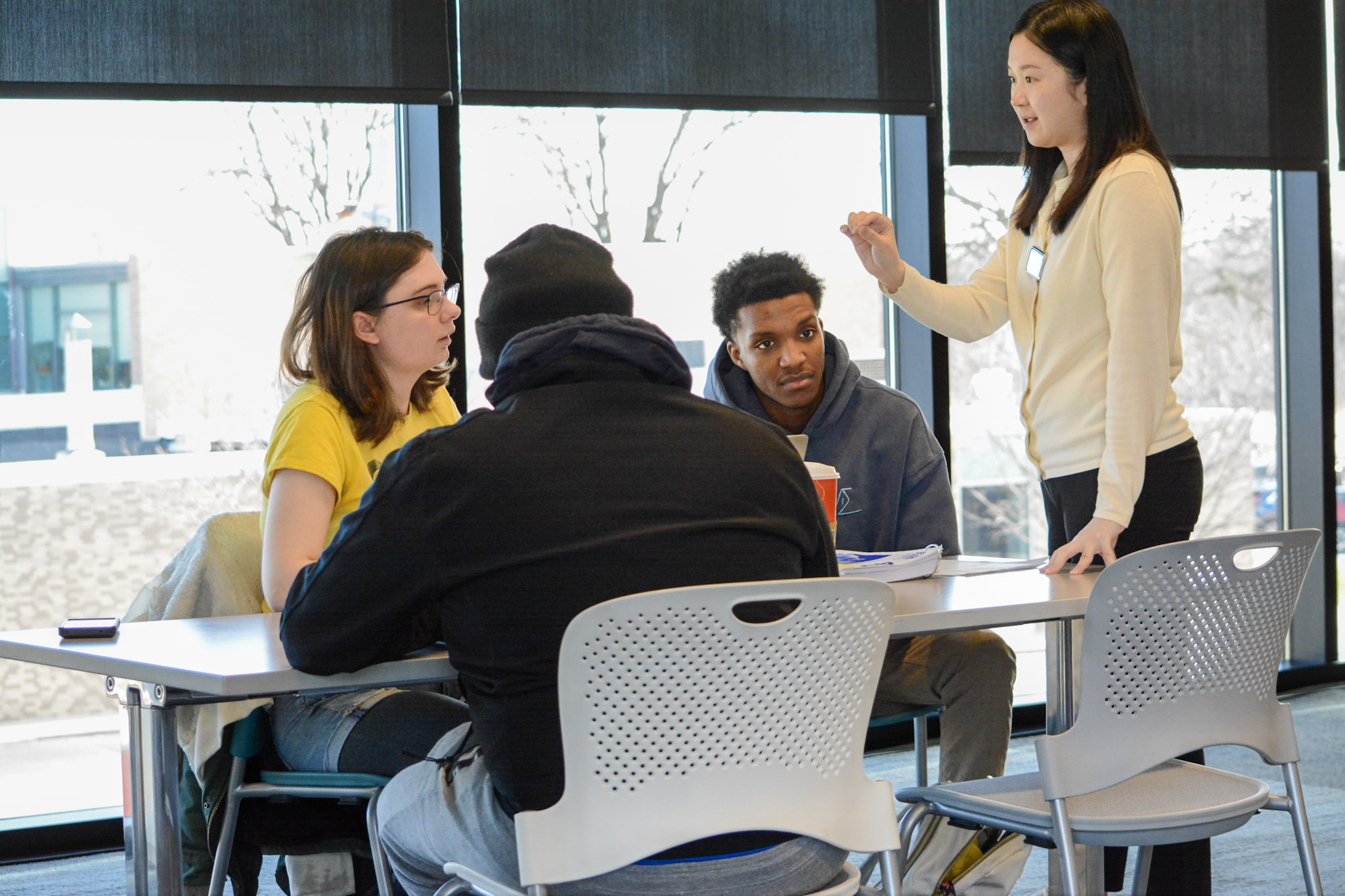 An instructor stands and speaks to three seated individuals. They are sitting at a table in a bright, modern space with large windows.
