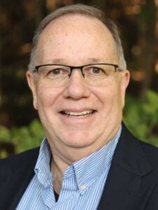 John Waldo smiles while wearing a blue blazer, light-blue, striped shirt and glasses. He has grayish-brown hair and brown eyes.