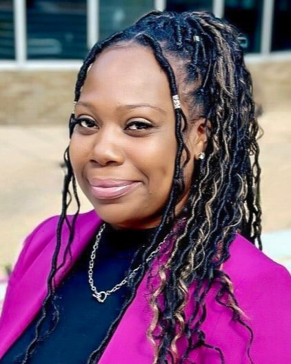 A woman with long, braided hair smiles while standing outdoors. She is wearing a pink blazer, black shirt and a necklace.