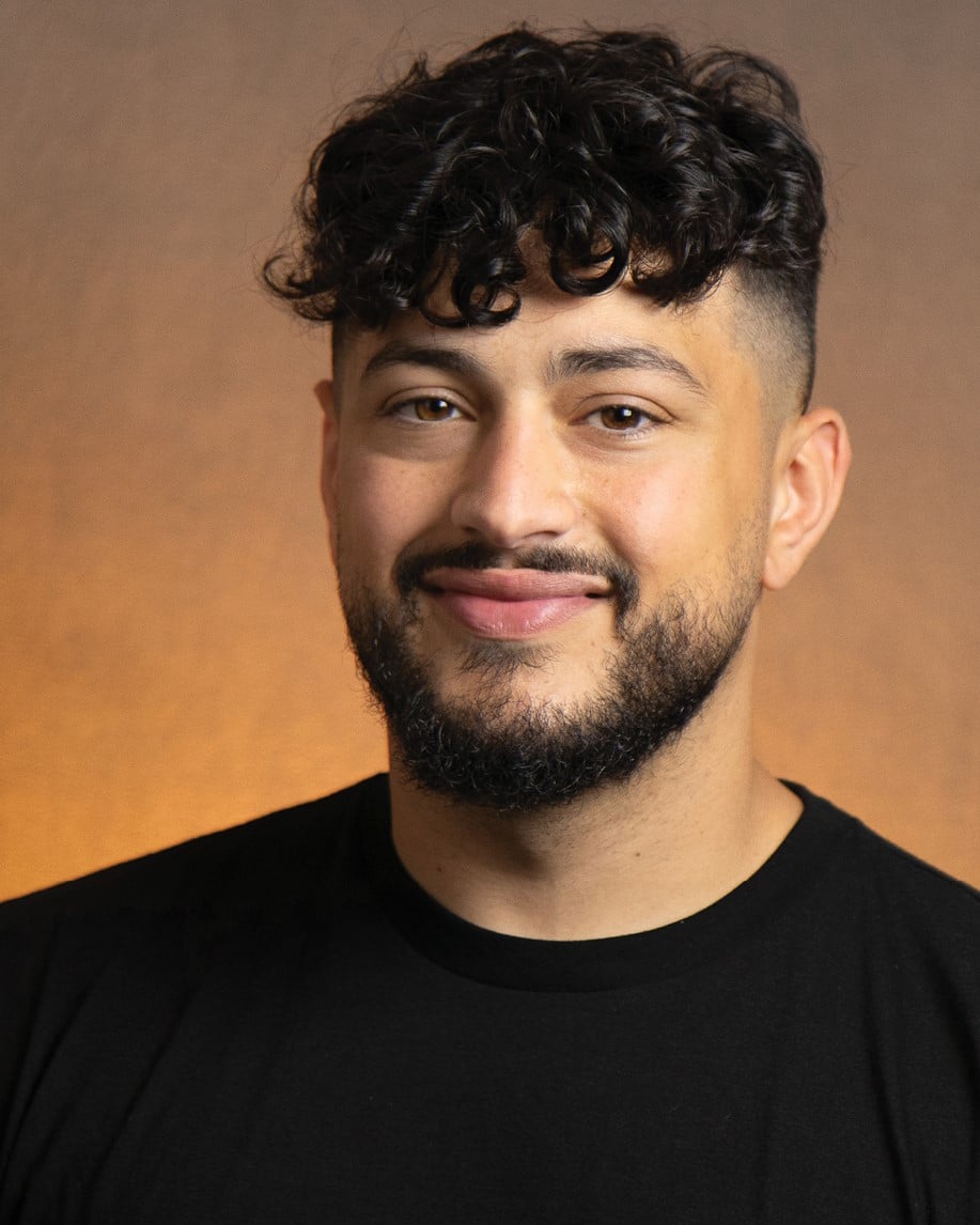 A man with curly hair and a dark beard smiles for a professional portrait. The background is a softly lit, orange color.