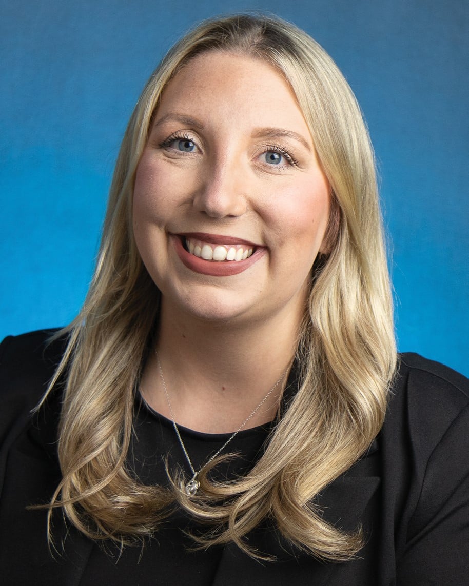 A woman with long blonde hair smiles for a professional portrait. The background is a vibrant blue color.