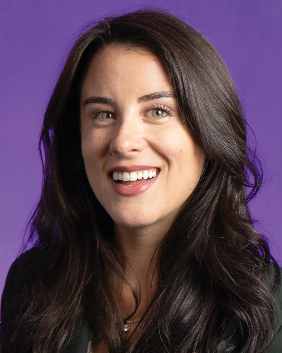 A woman with long brunette hair smiles for a professional portrait. The background is a vibrant purple color.