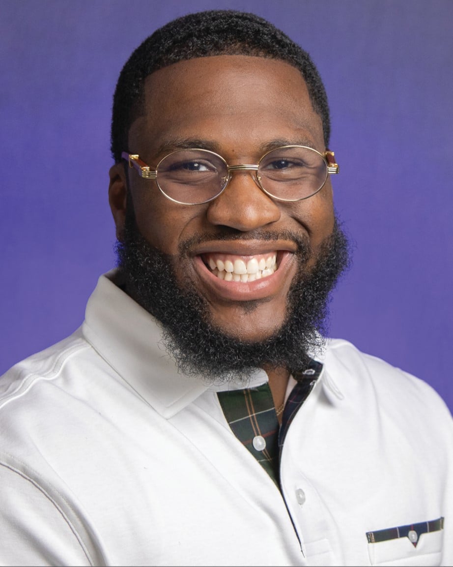 A man with glasses smiles broadly for a professional portrait. The background is a soft purple color.