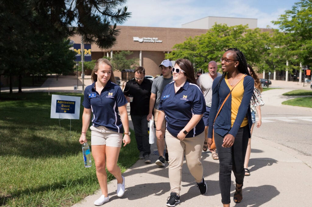 A group of people walking together on a campus path, with a sign for an "Admissions Event" visible nearby.