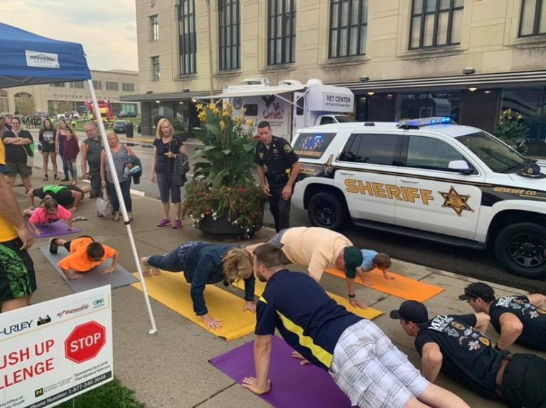 A group of people participating in a push-up challenge on a street, with a sheriff's vehicle and onlookers in the background.