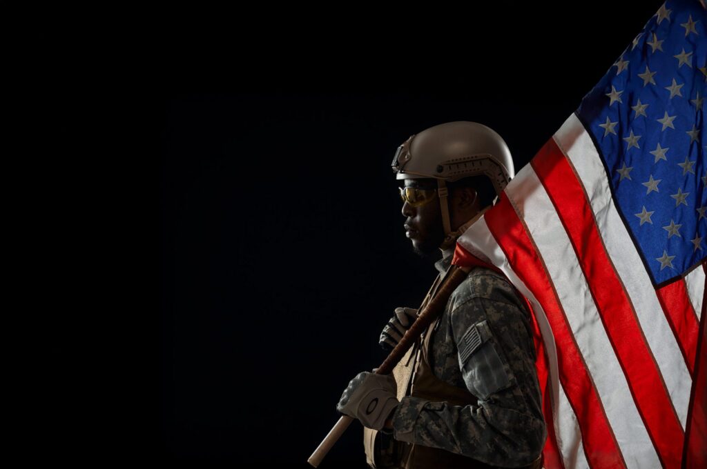A person in military attire stands in profile, holding an American flag against a dark background.