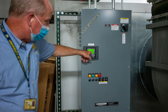 A person points at a control panel with various buttons and a display in an industrial setting.