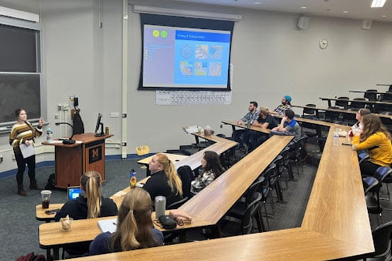 A person presents in a classroom with a projector screen displaying images, while several students are seated at desks.