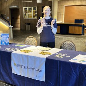 A person stands behind a table displaying a "Planet Blue Ambassador" banner, holding two bananas.