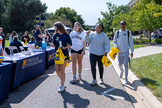 A group of four people walking together outdoors, holding bags, with a table displaying promotional materials in the background.