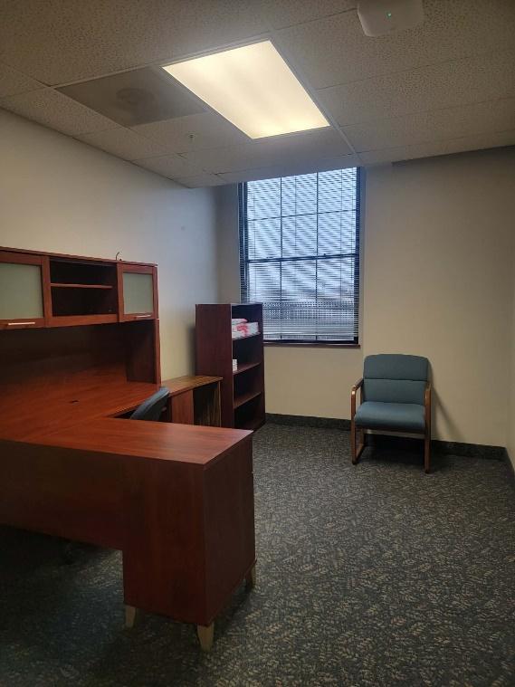 Office room with a wooden L-shaped desk, a blue cushioned chair, a small bookshelf, and a window with blinds.