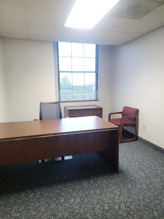 Office room with a wooden desk, a mesh office chair, a small wooden cabinet under a window with blinds, and a red cushioned guest chair.