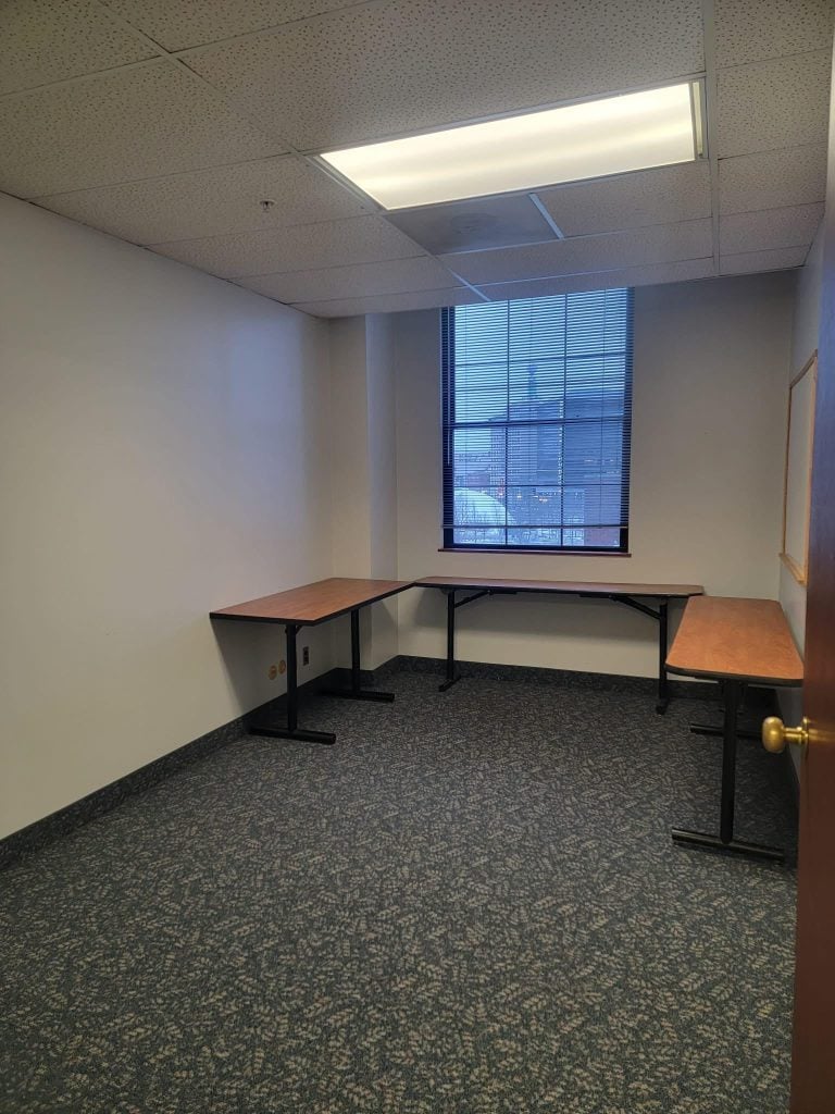 Empty office room with three wooden tables arranged in an L shape under a window with blinds, carpeted floor, and fluorescent ceiling light.