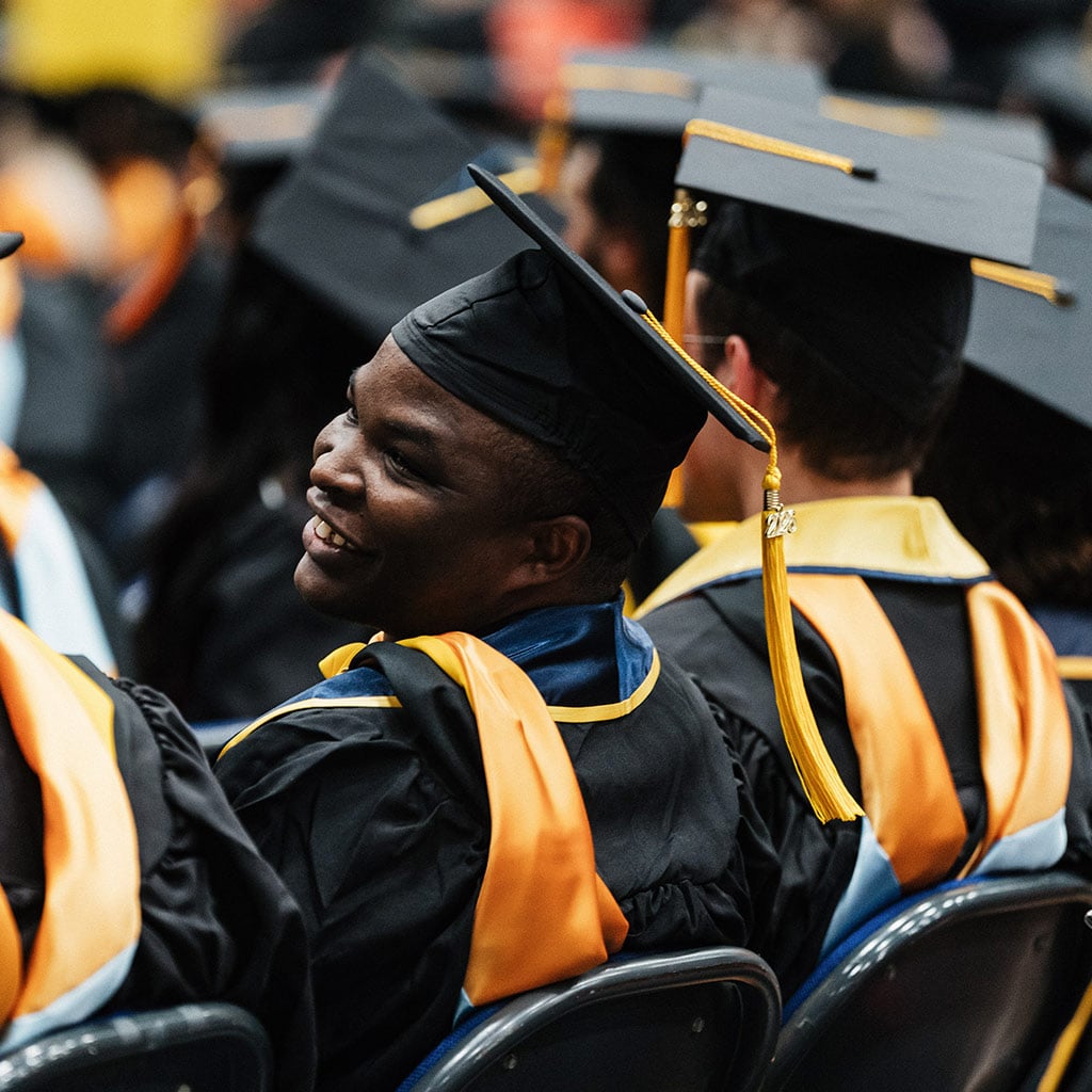 A group of graduates seated in caps and gowns, with one person's face obscured, attending a graduation ceremony.