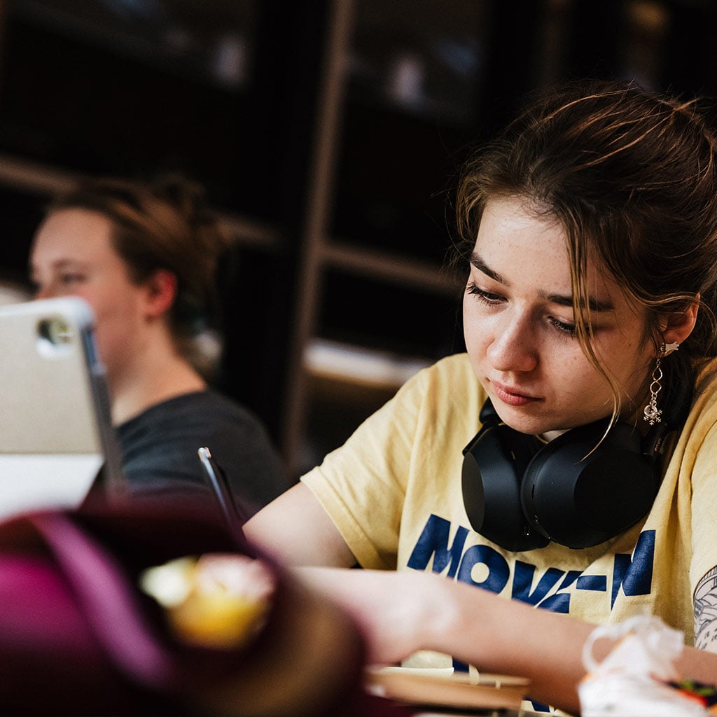 A person wearing headphones is writing on a notebook at a table, with another person visible in the background.