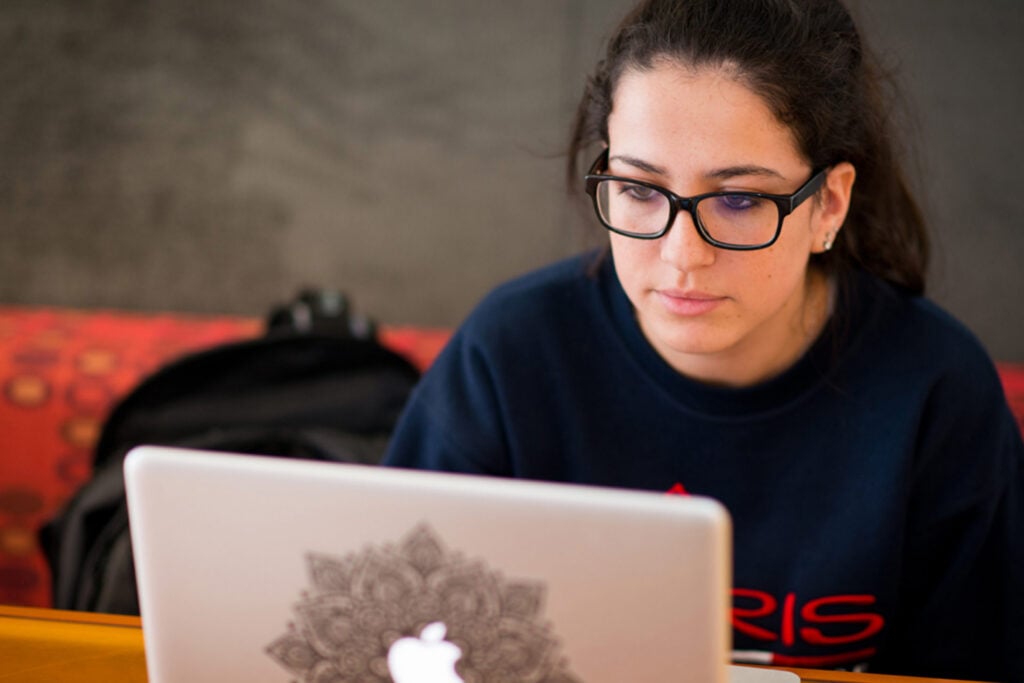 A person wearing a dark sweatshirt with "PARIS" printed on it is sitting in front of a laptop with a decorative sticker on the lid.