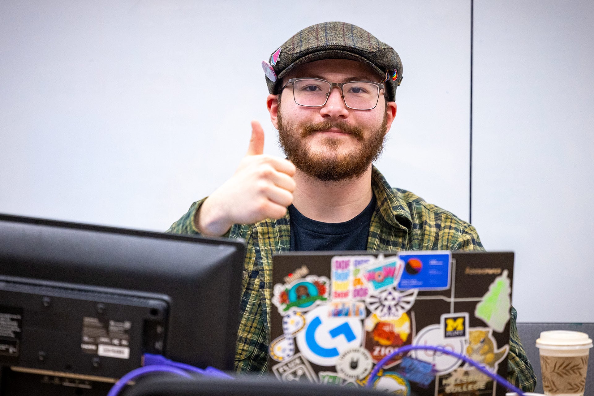 A person gives a thumbs-up while sitting at a desk with a laptop covered in stickers and a coffee cup nearby.