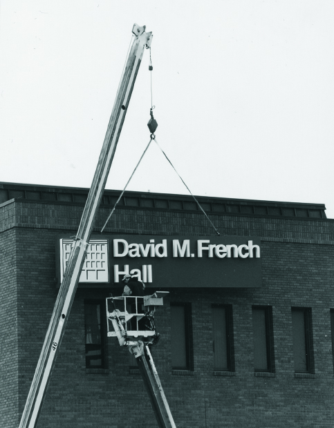 A crane installing signage reading ‘David M. French Hall’ on a campus building.