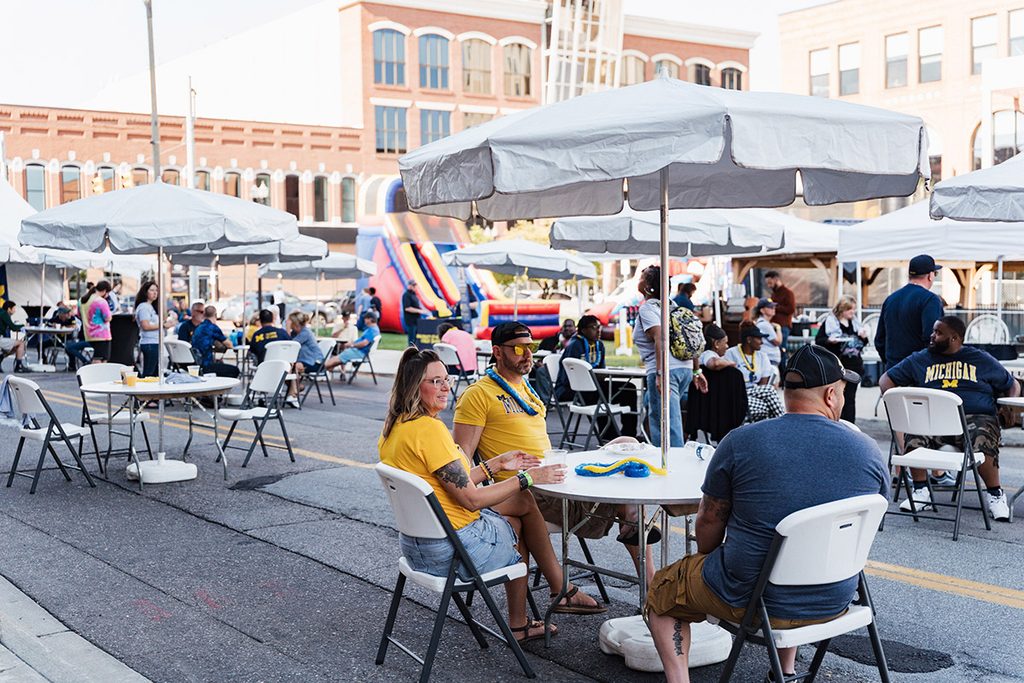 A group of people sitting at tables under white tents in a street setting, with a bounce house visible in the background.