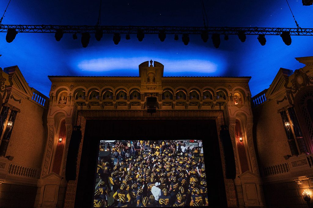 A theater interior with a large screen displaying a crowd of people, under a blue ceiling with decorative architecture.