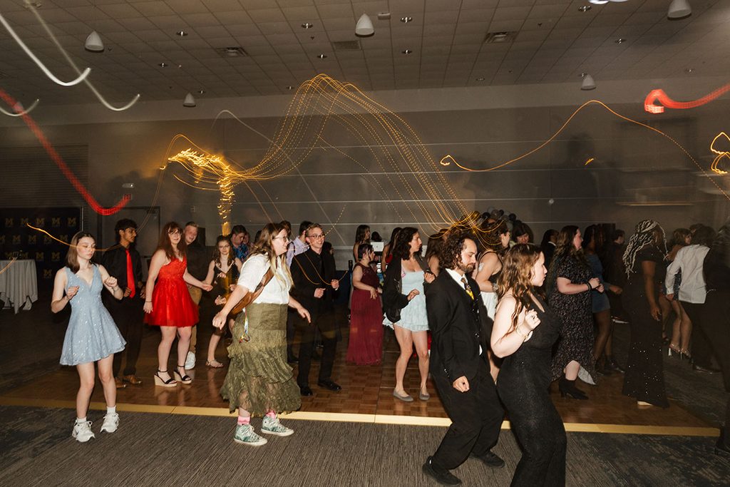 A group of people dance at a lively event, with colorful lights and a festive atmosphere in a large indoor space.