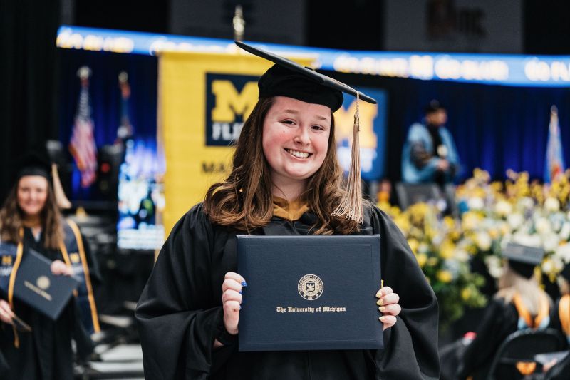 Abigail Weycker in a graduation cap and gown smiles while holding a diploma from the University of Michigan at a graduation ceremony.