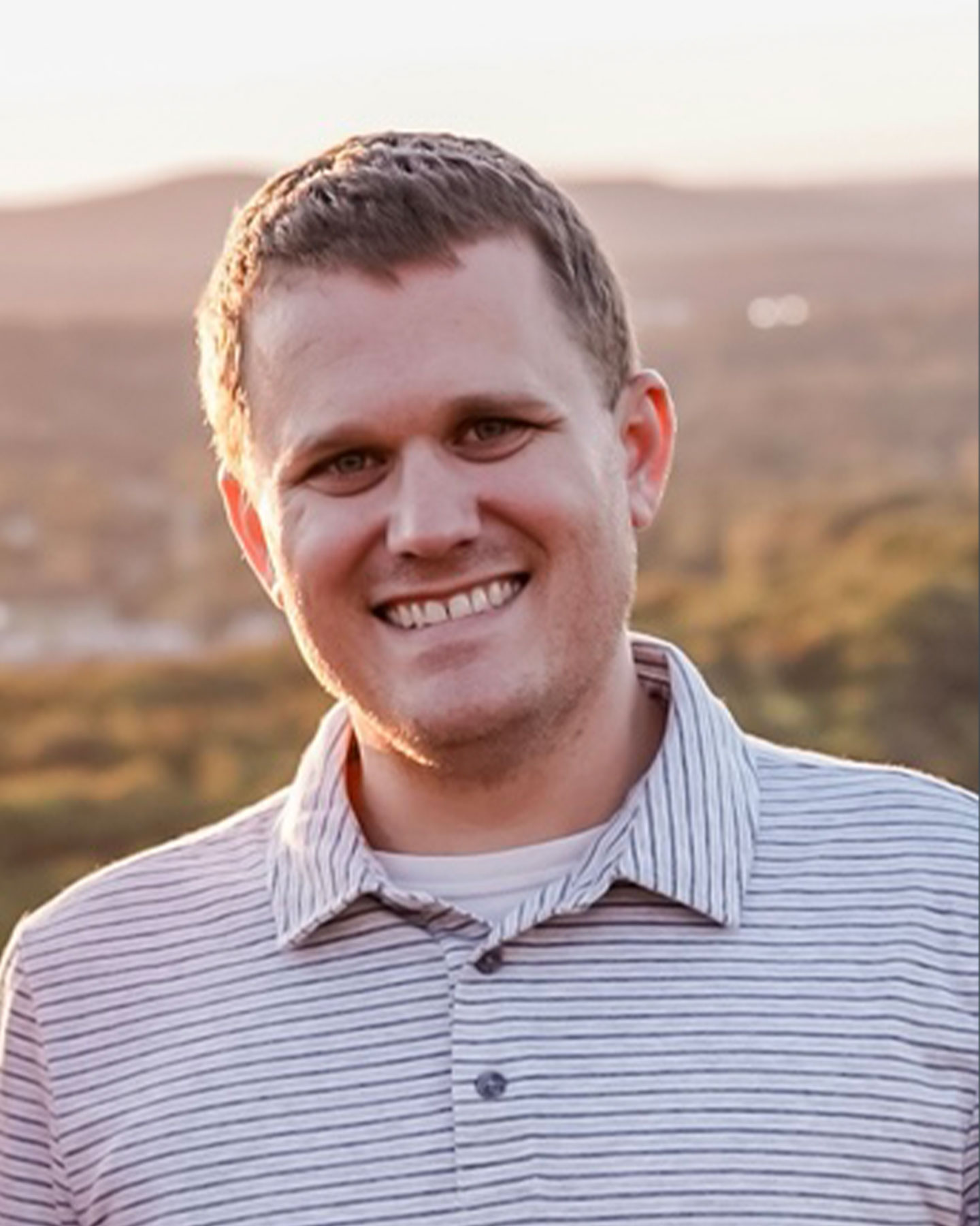 Kyle Cantrell smiles while standing outdoors with a blurred landscape in the background during sunset.